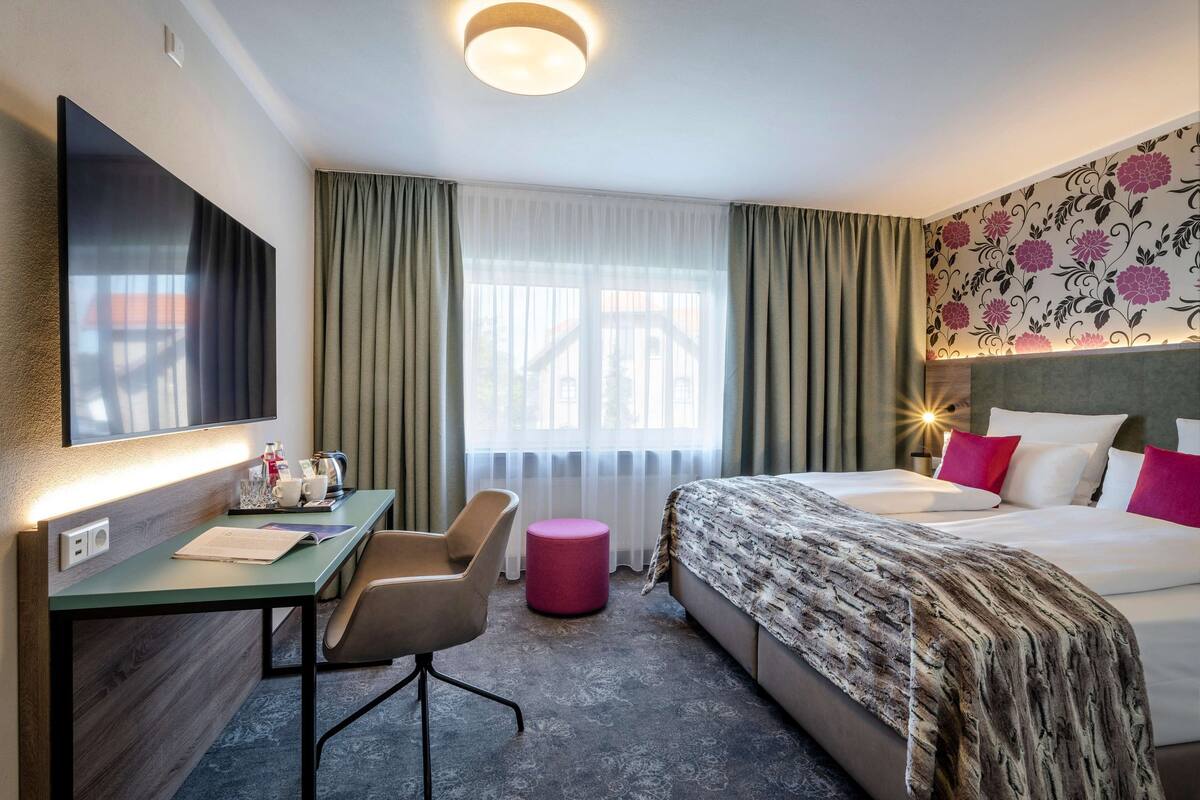 Habitación de hotel con cama, escritorio con silla, televisor, ventana con cortinas y pared con estampado floral.