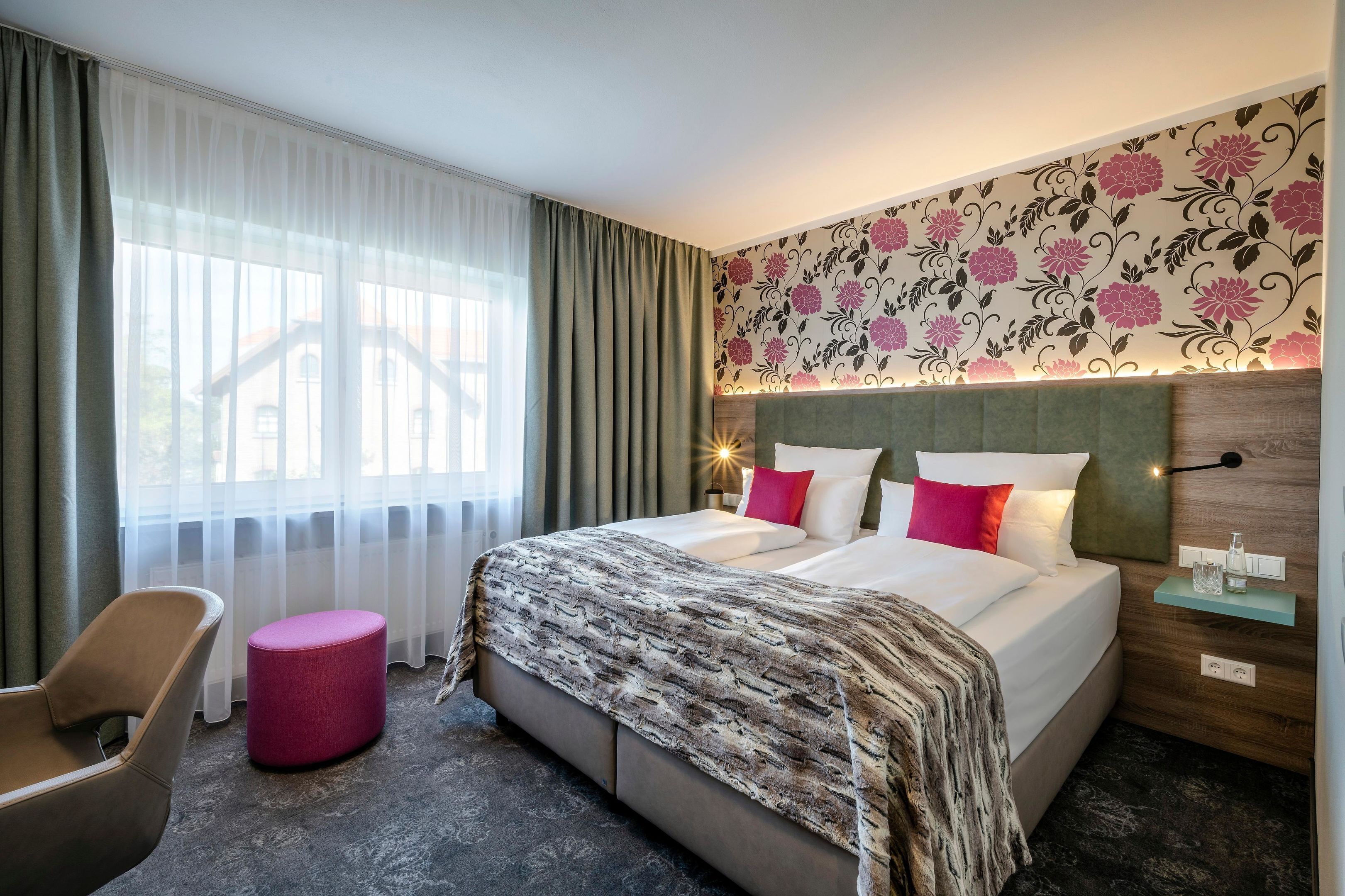 Habitación de hotel con una cama, una silla, un otoman redondo, una mesita de noche y un ventanal con cortinas.