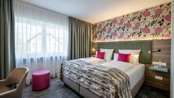 Habitación de hotel con una cama, una silla, un otoman redondo, una mesita de noche y un ventanal con cortinas.