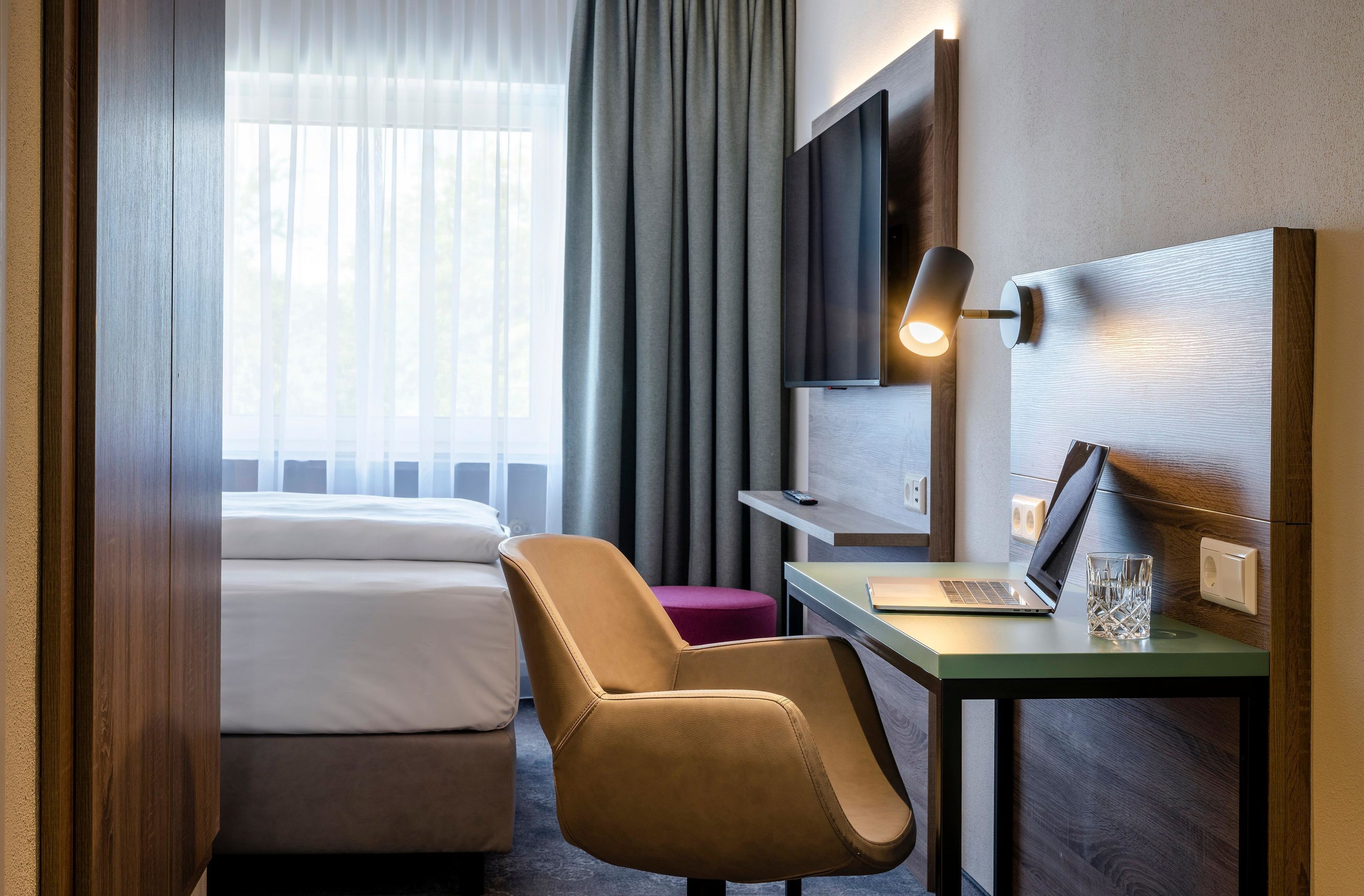 A hotel room with a bed, a desk with a laptop, a chair, a TV mounted on the wall, and a window with curtains.