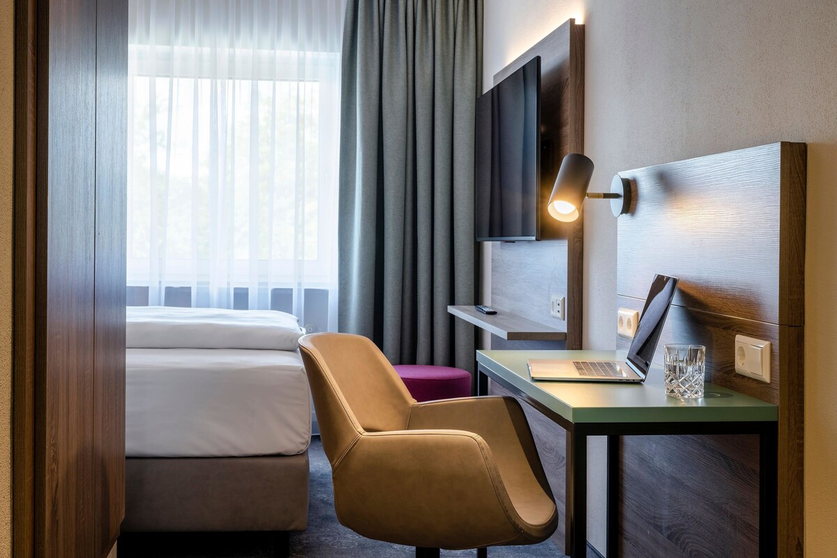 Une chambre d’hôtel avec un lit, un bureau avec un ordinateur portable, une chaise, un téléviseur fixé au mur et une fenêtre avec des rideaux.