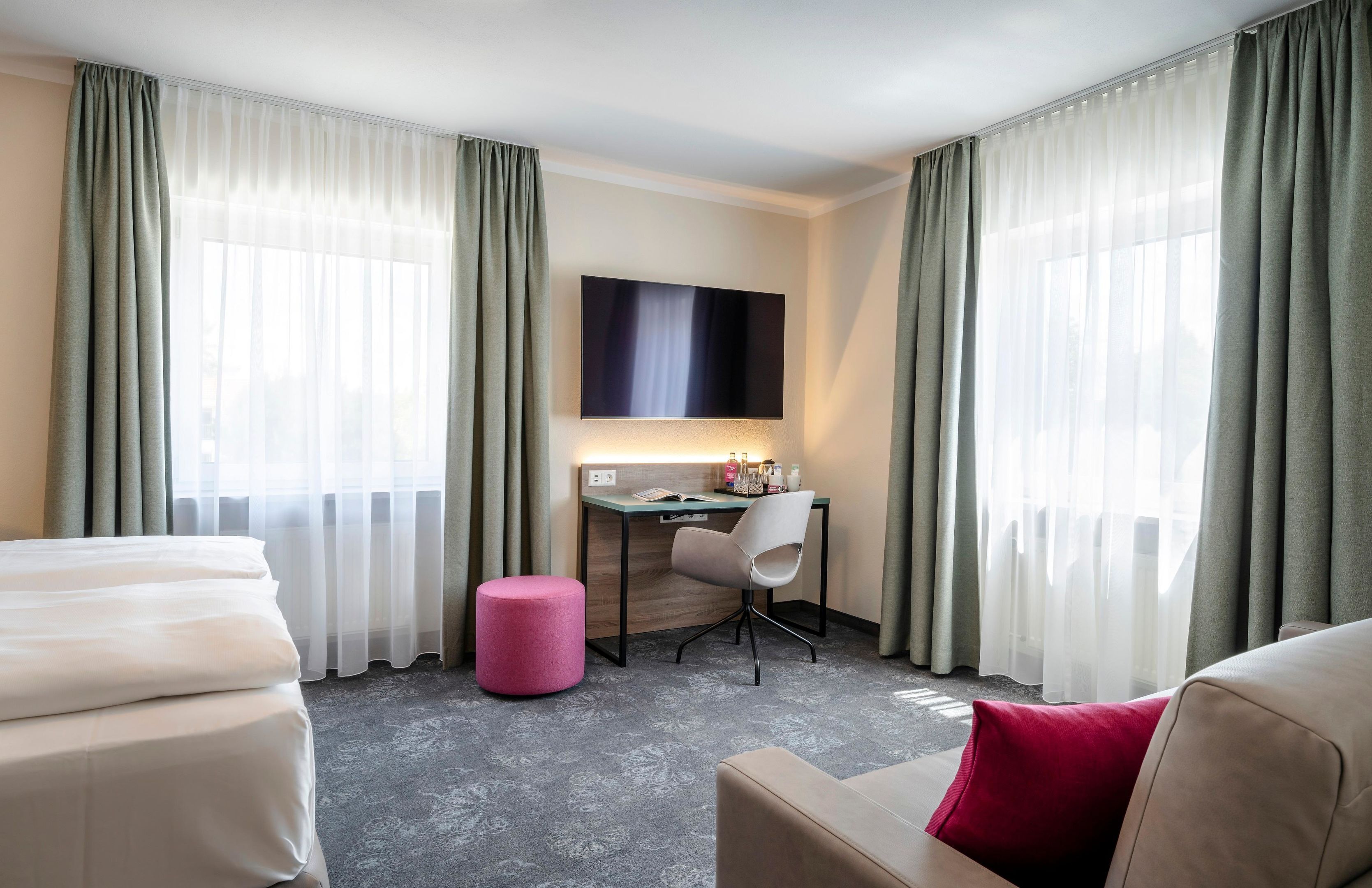Una habitación de hotel con una cama, un escritorio con una silla, un reposapiés rosa y un televisor montado en la pared.