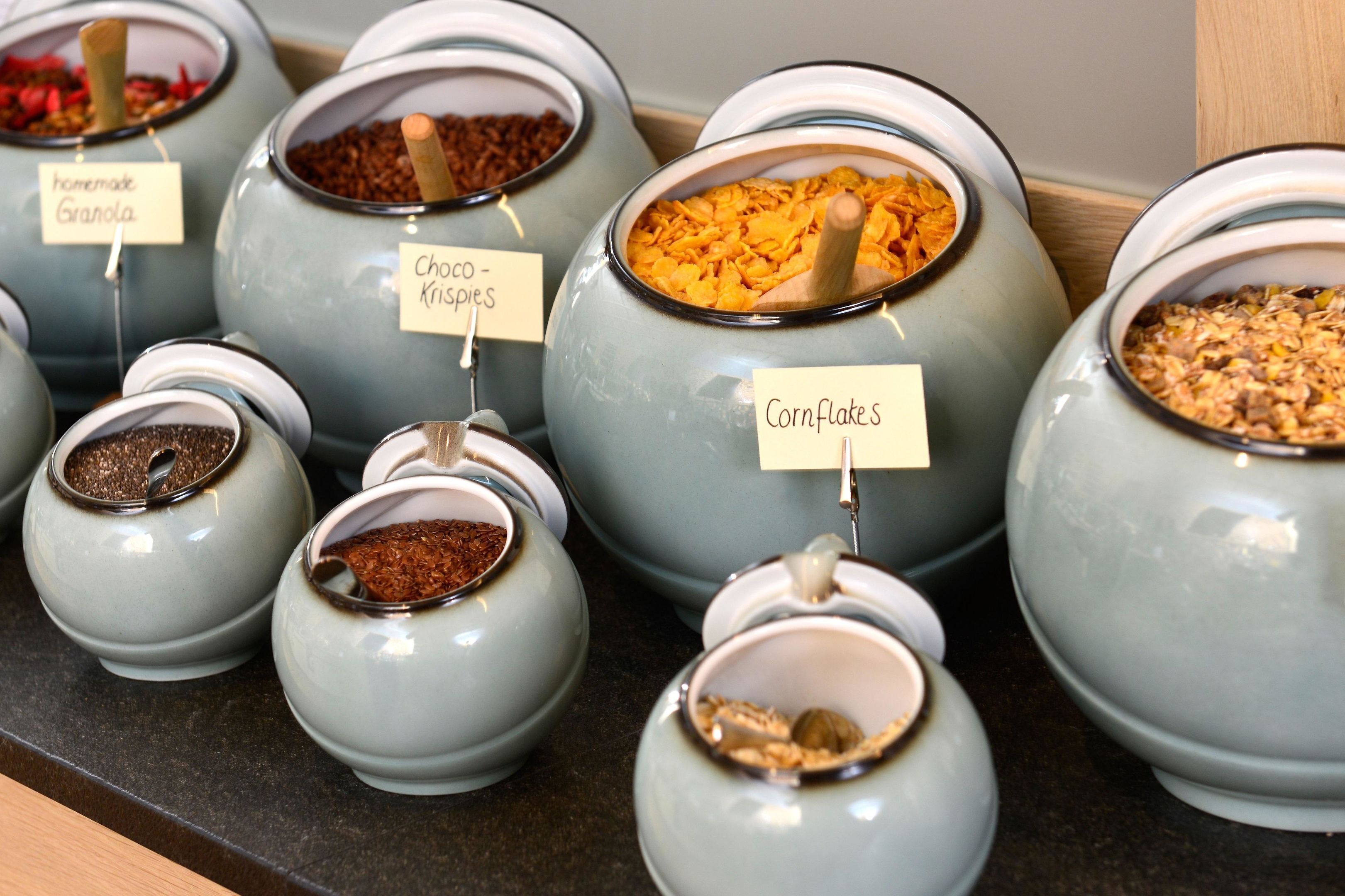 On trouve une variété d’aliments dans des récipients en céramique, chacun étant muni d’une étiquette. Parmi ces aliments, on peut citer du granola fait maison, des graines de chia, des biscuits aux graines de chia et des céréales de maïs.