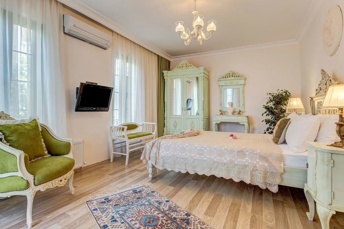 Un dormitorio con cama, televisor, sillón verde, silla blanca, armario verde y mesita de noche con lámpara.