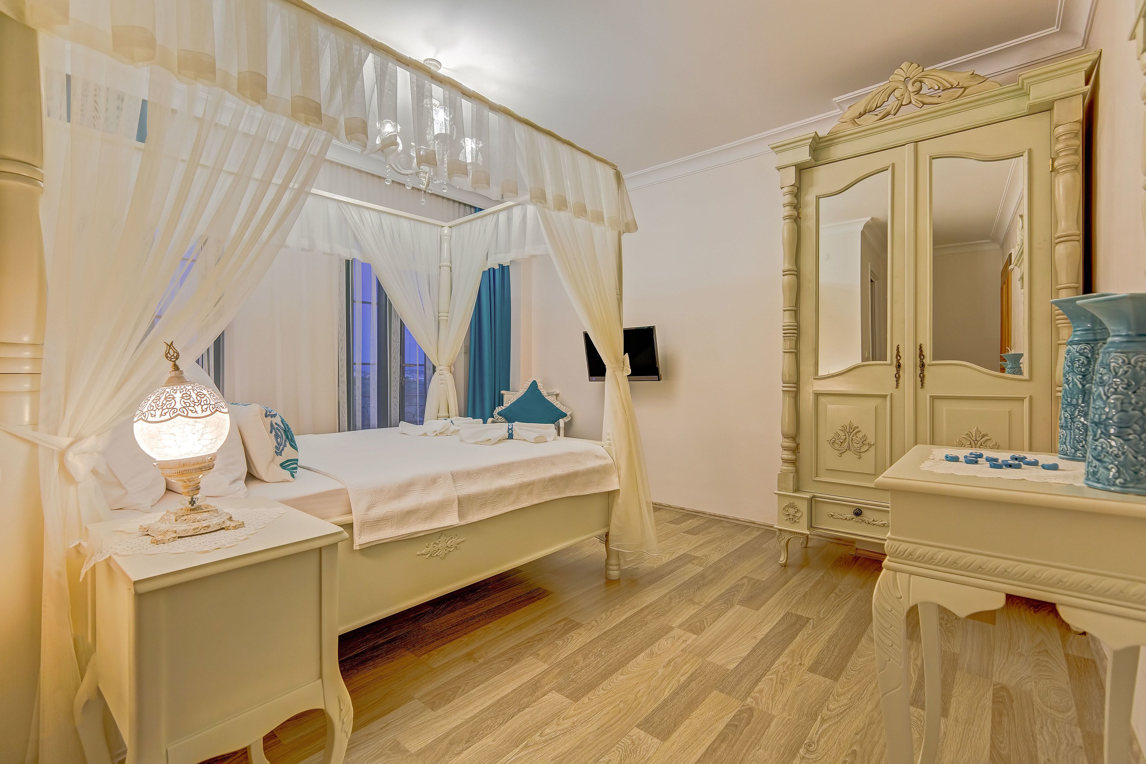 A bedroom with a four-poster bed, a nightstand with a lamp, a dresser with a mirror, and a blue vase.