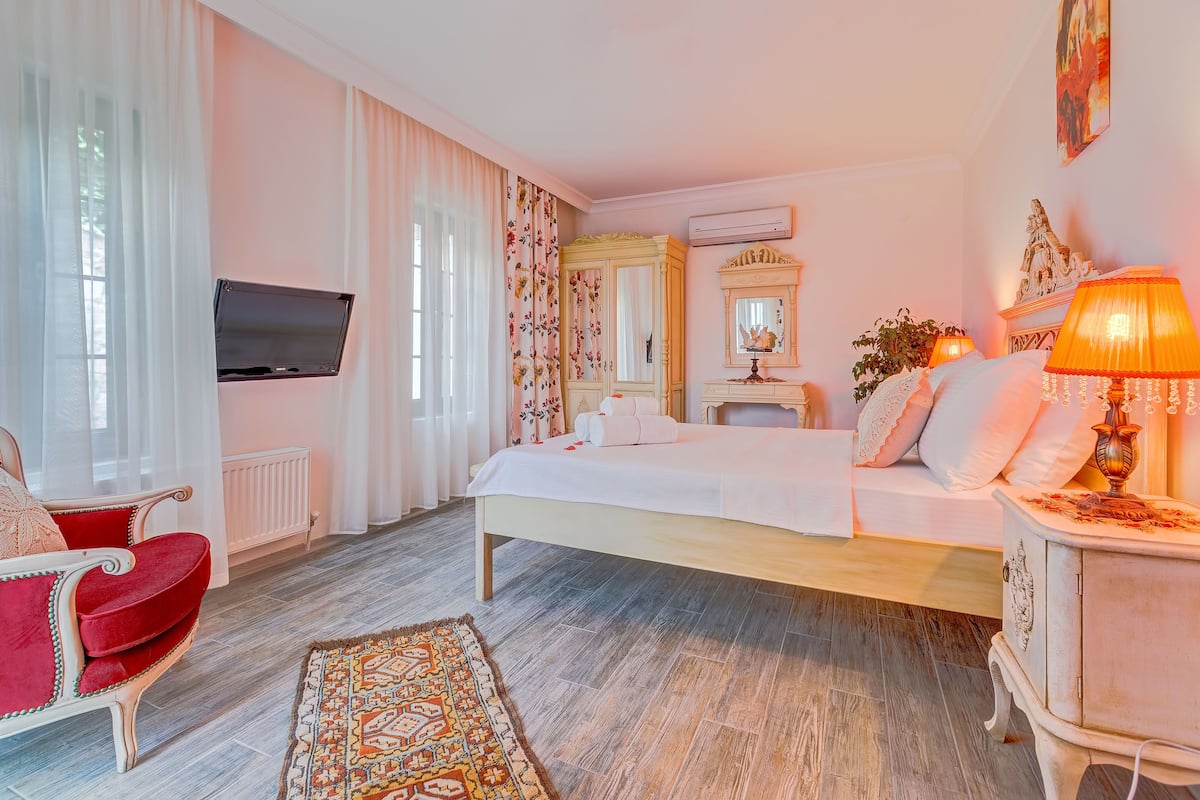 Un dormitorio con una cama, un televisor, una silla roja, una mesita de noche con lámpara y un espejo decorativo.