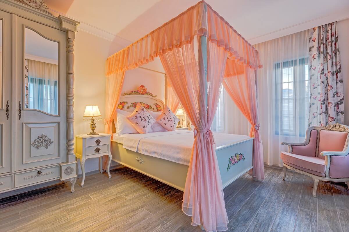 Una cama con dosel y cortinas florales, un aparador clásico y un sillón rosa.