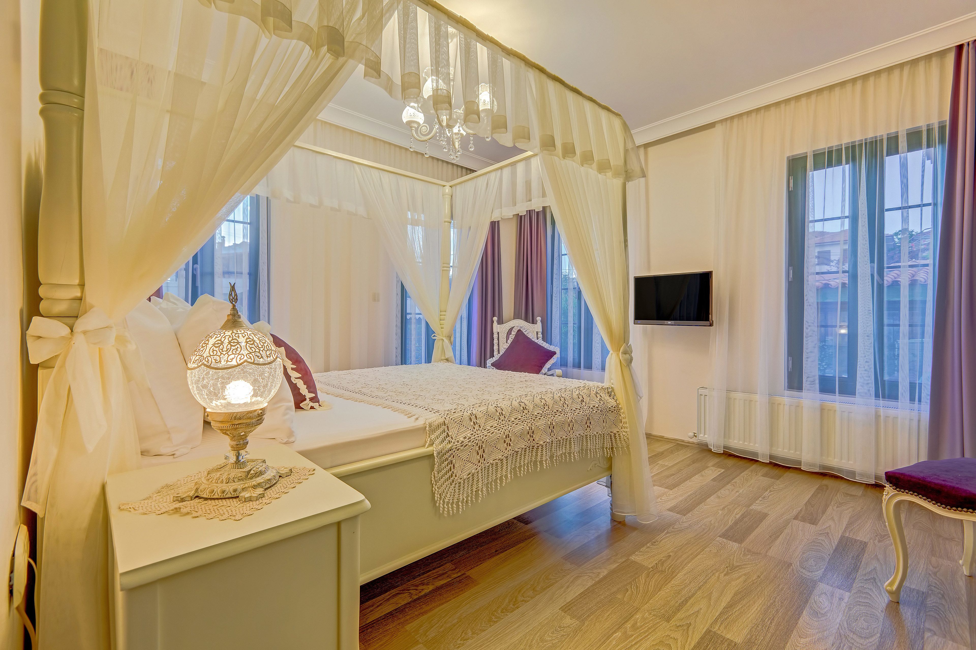 A bedroom with a four-poster bed, a bedside lamp, a TV, and a view of the outside through large windows.