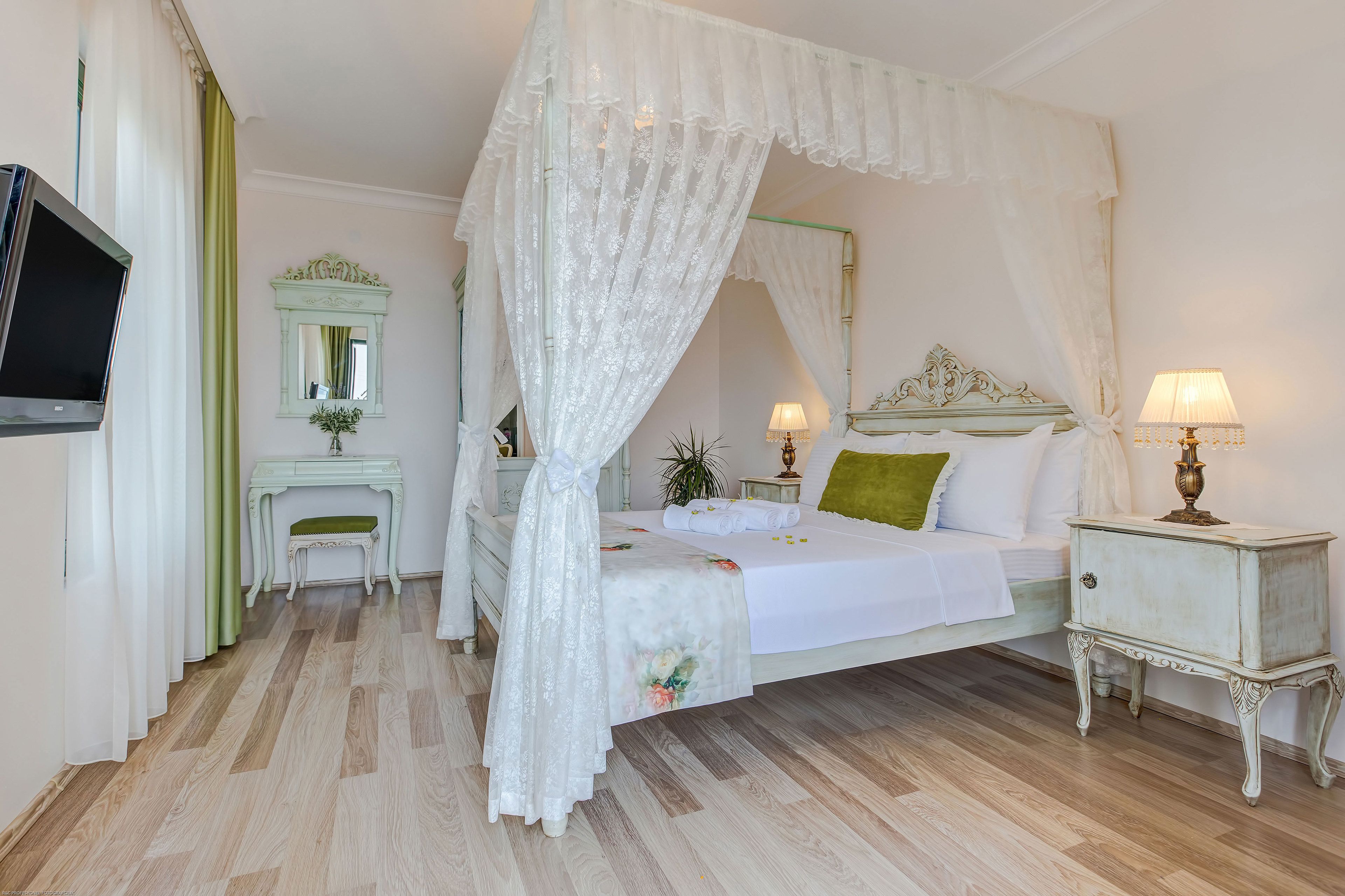 Ein Schlafzimmer mit Baldachinbett, Nachttisch mit Lampe, kleiner Schreibtisch mit Stuhl, Spiegel und Fernseher an der Wand.
