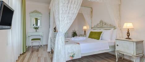 A bedroom with a canopy bed, a bedside table with a lamp, a small desk with a chair, a mirror, and a television on the wall.