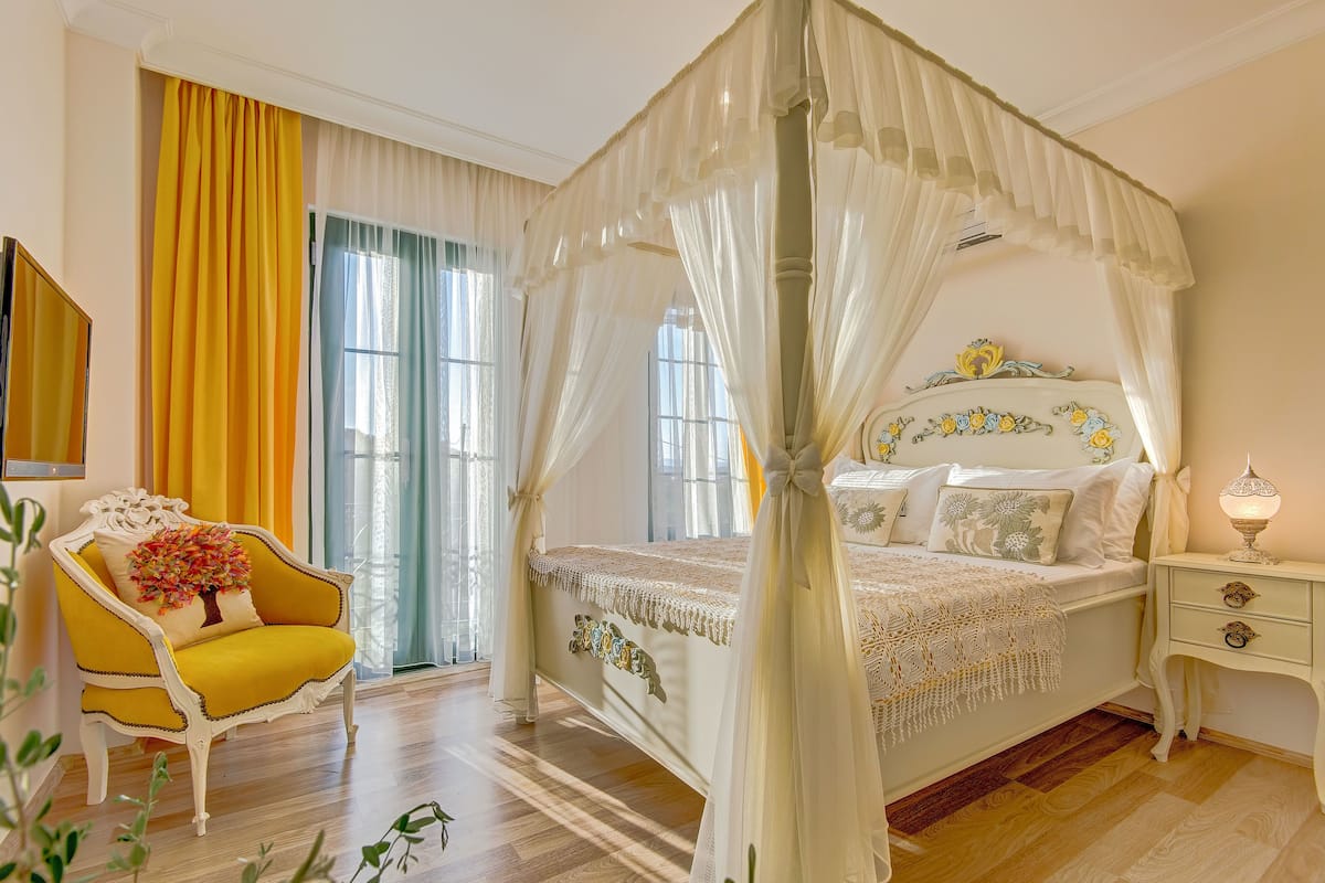 Una cama con dosel y cuatro columnas, con una paleta de colores blanco y dorado, un sillón amarillo con un cojín floral y una mesita de noche con una lámpara.