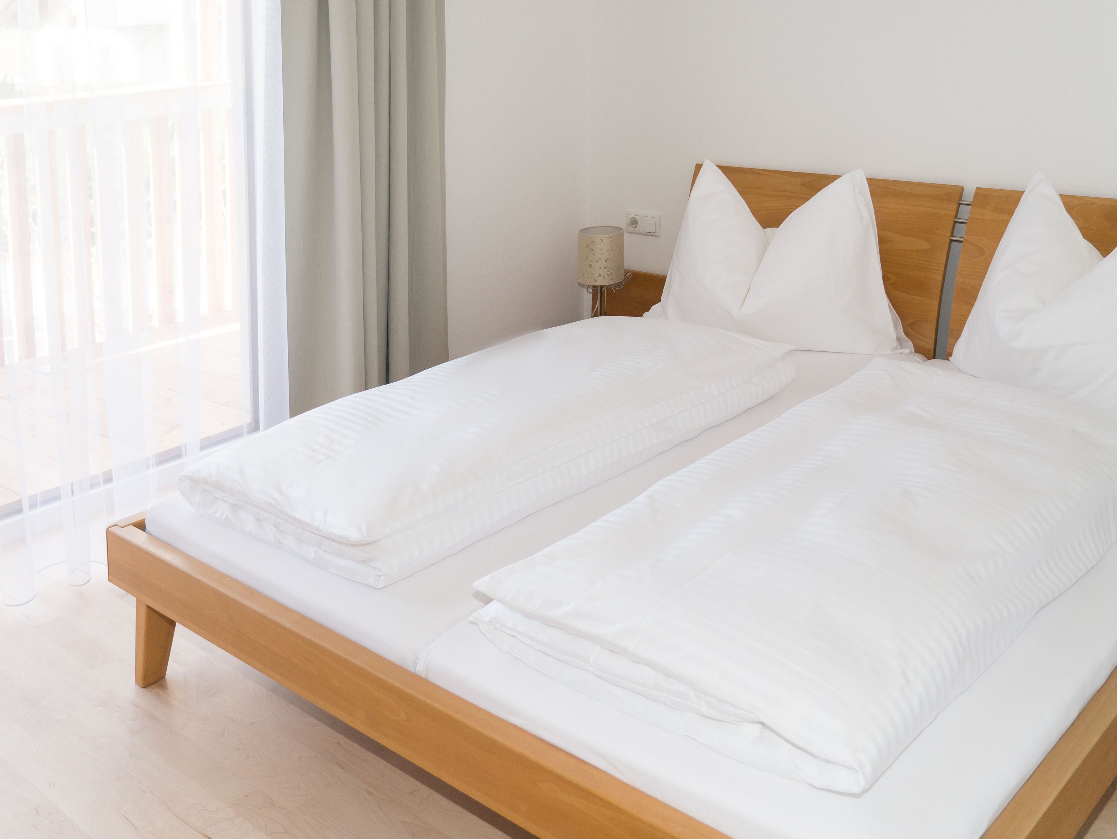 A double bed with wooden headboard and white bedding in a bright room with a window and curtains.