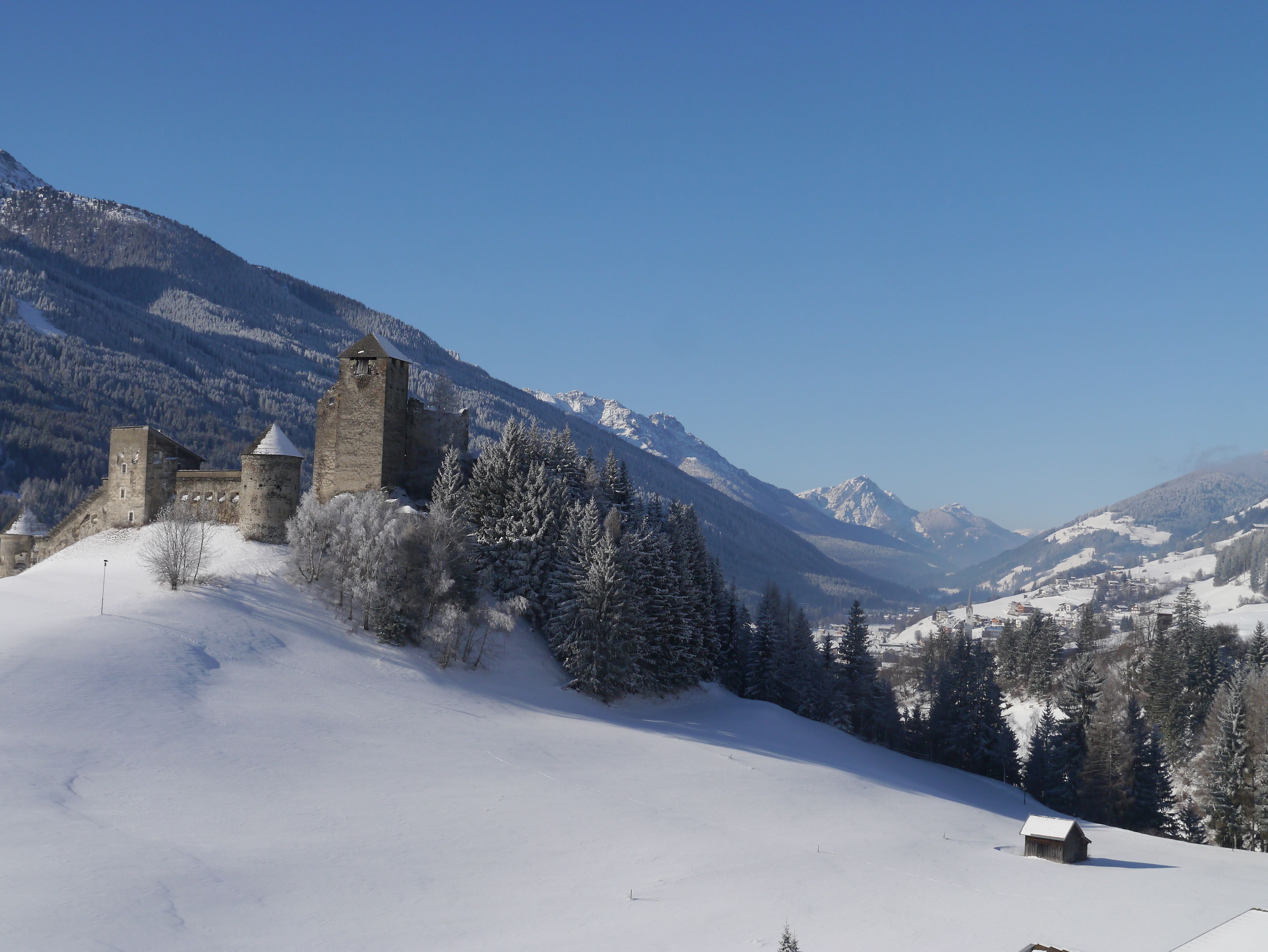 A snow-covered landscape with a historic stone building featuring towers, surrounded by snow-laden trees and mountains in the background.