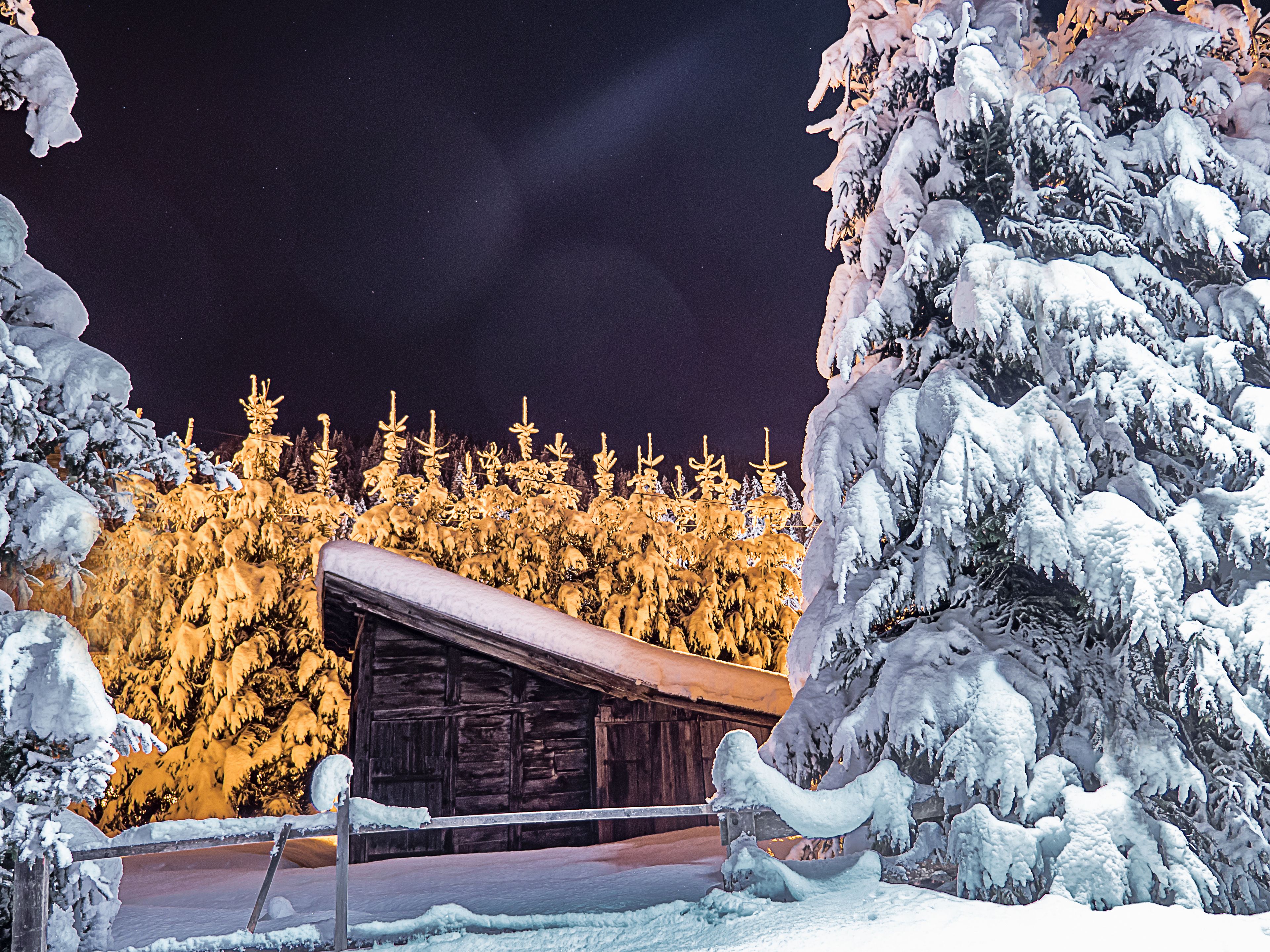 Lumipeitteinen maisema, jossa on puinen mökki ja taustalla metsä.
