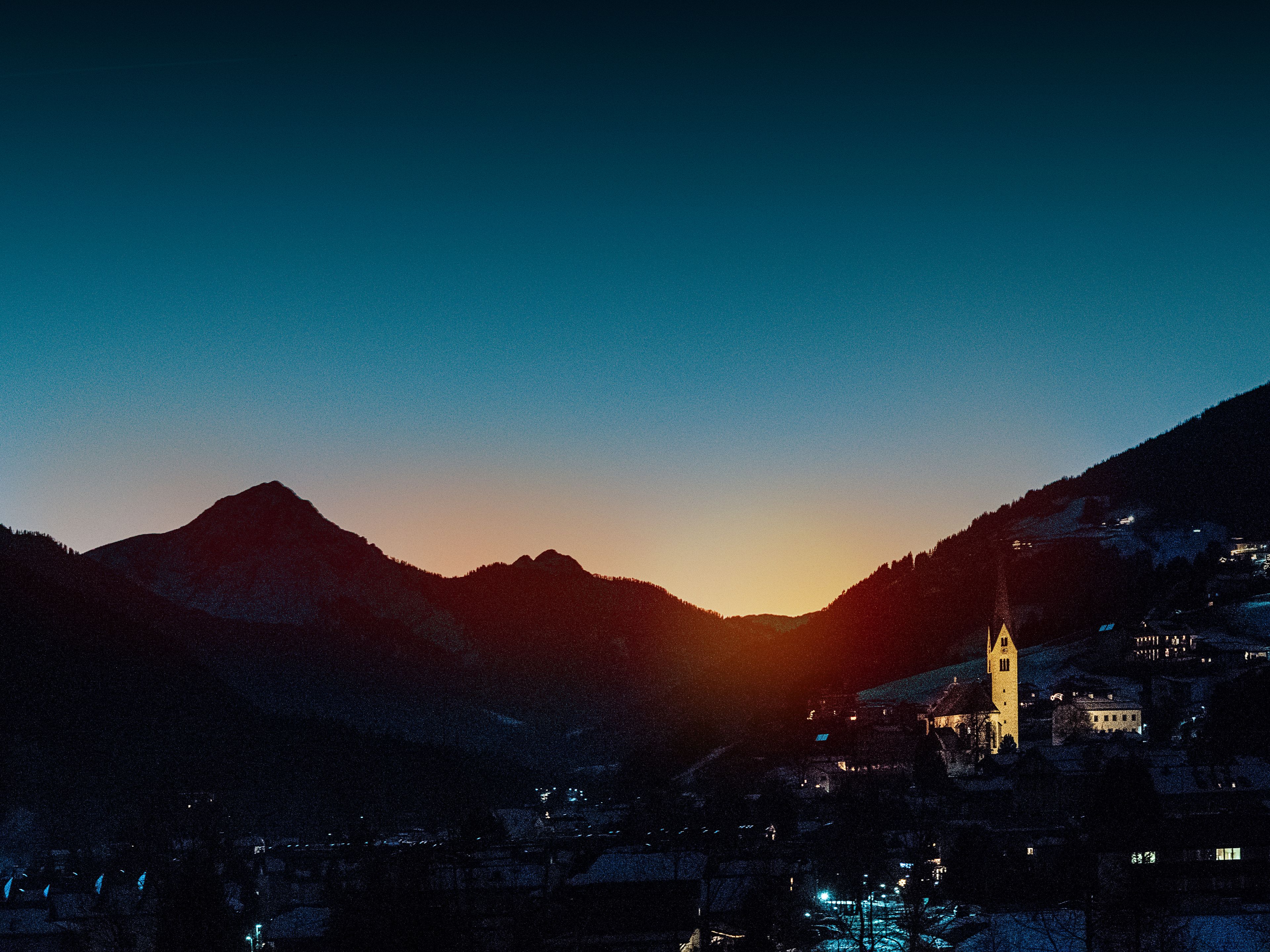 A town with a prominent church tower, surrounded by mountains and lit buildings.