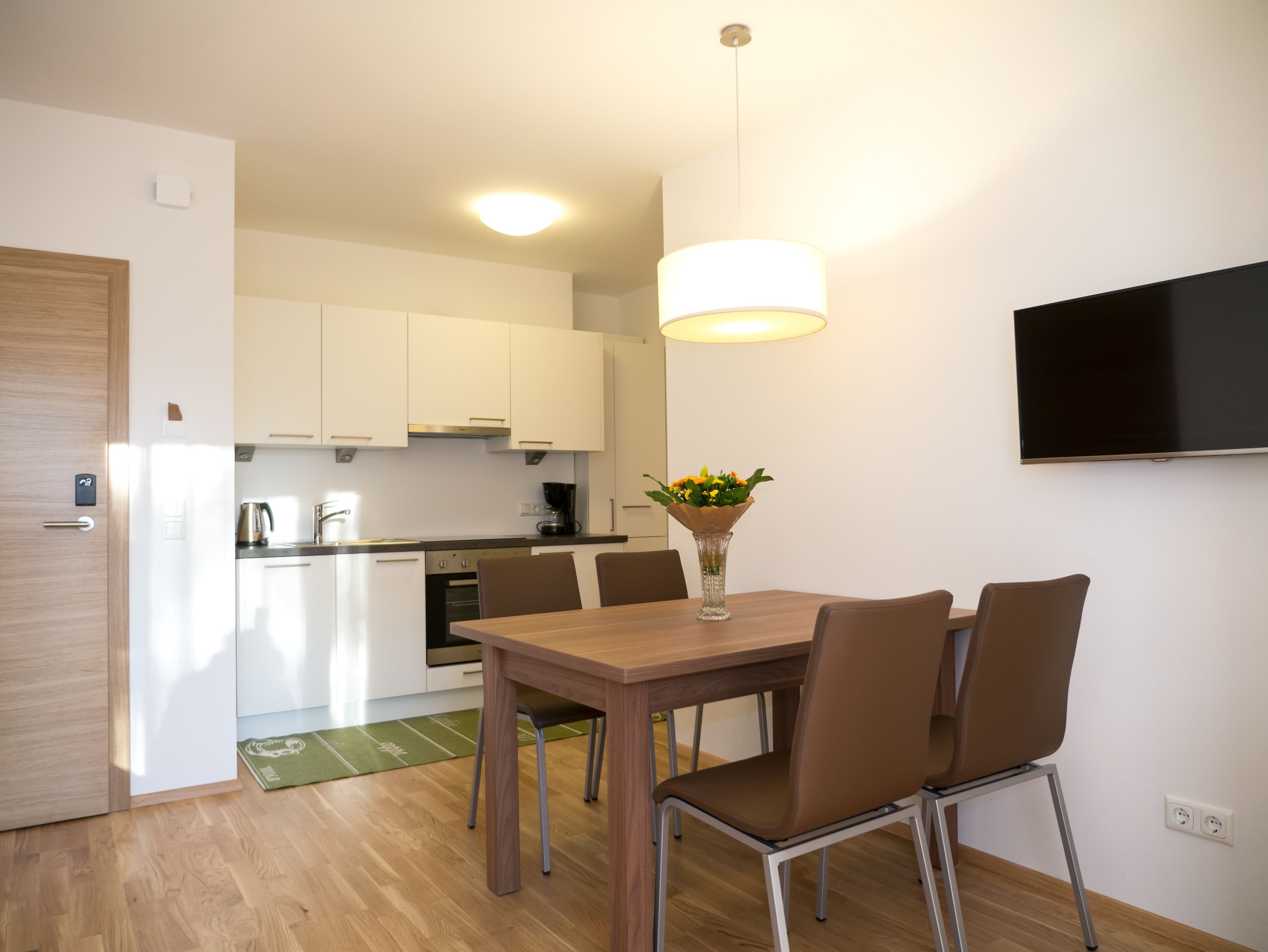 Een moderne keuken met witte kasten, een eettafel met stoelen, een vaas met bloemen en een flatscreen televisie aan de muur.