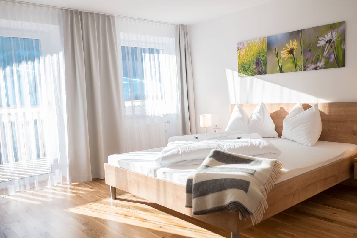 Una habitación de hotel moderna con una cama grande, suelo de madera y un ventanal con cortinas traslúcidas.