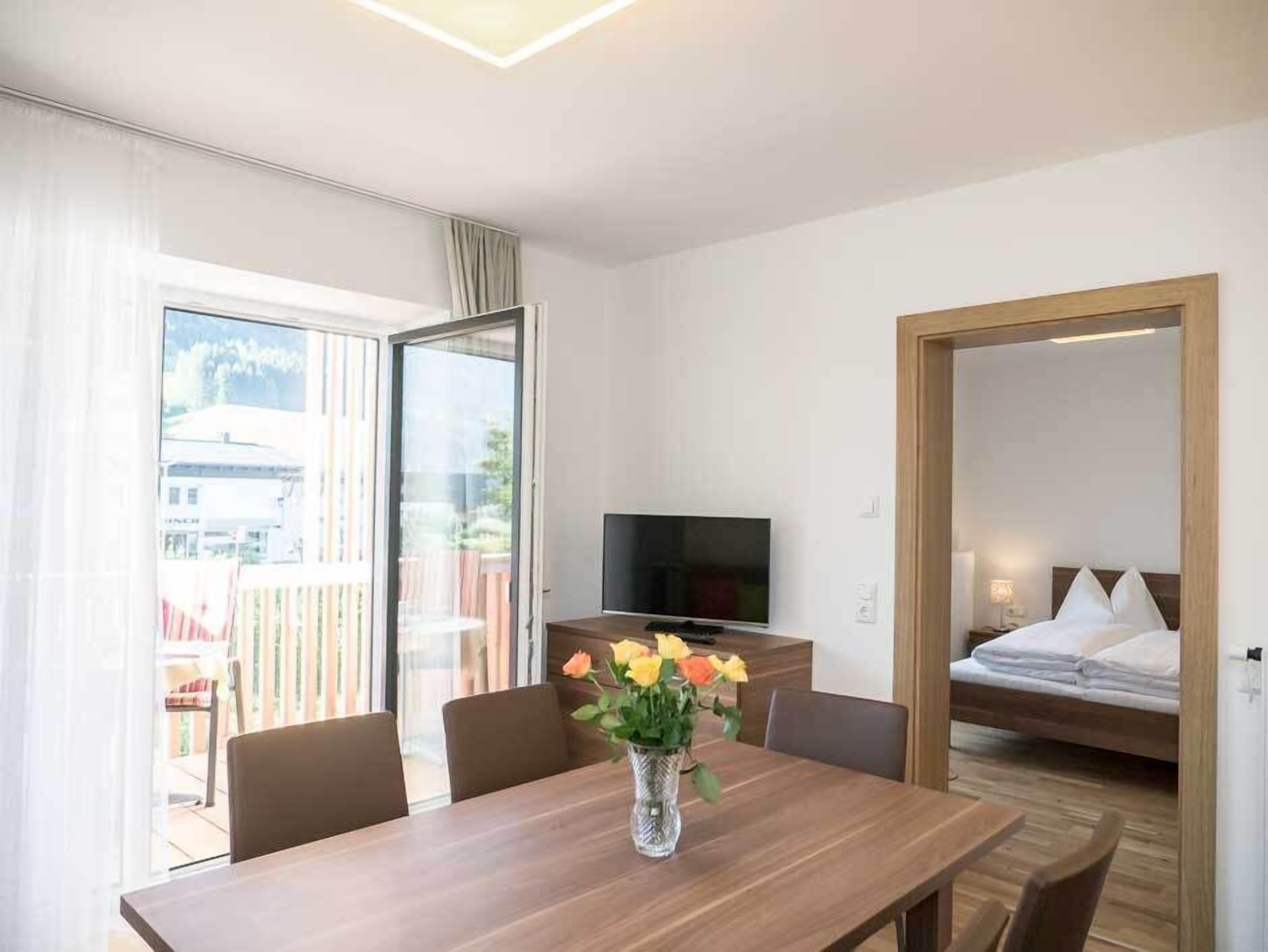 Habitación de hotel con una mesa de comedor de madera, televisor, cama y balcón con vista a edificios.