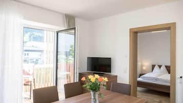 Habitación de hotel con una mesa de comedor de madera, televisor, cama y balcón con vista a edificios.
