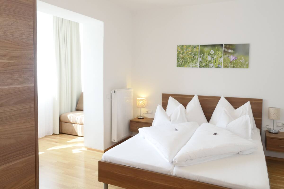 Una cama bien hecha con sábanas y almohadas blancas, cabecera de madera, mesita de noche con lámpara y radiador.