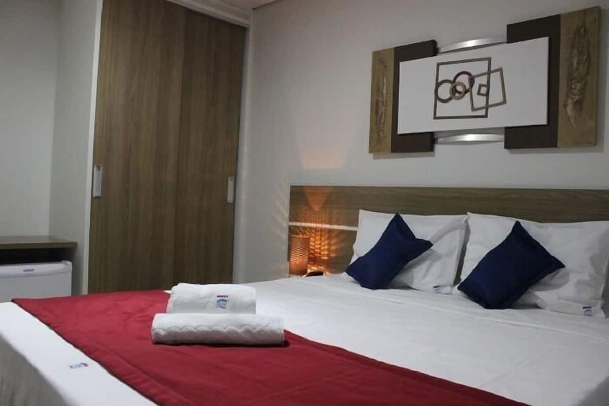 Een hotelkamer met een bed, een rode deken, twee kussens, een hoofdeinde, een aan de muur hangend kunstwerk en een houten deur.