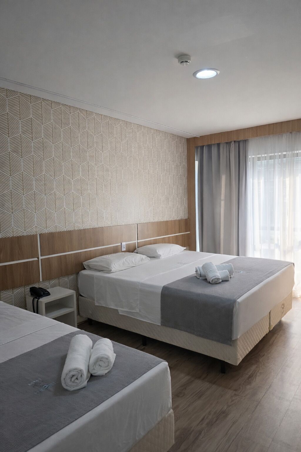 Une chambre d’hôtel avec deux lits, un mur orné d’un motif, une tête de lit en bois, une table de chevet avec un téléphone et une fenêtre avec des rideaux.