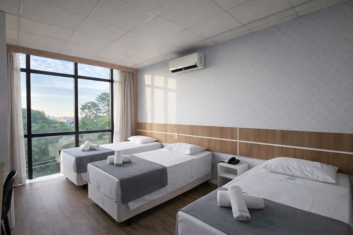 Een hotelkamer met drie bedden, een groot raam, een airconditioning en uitzicht op groen.