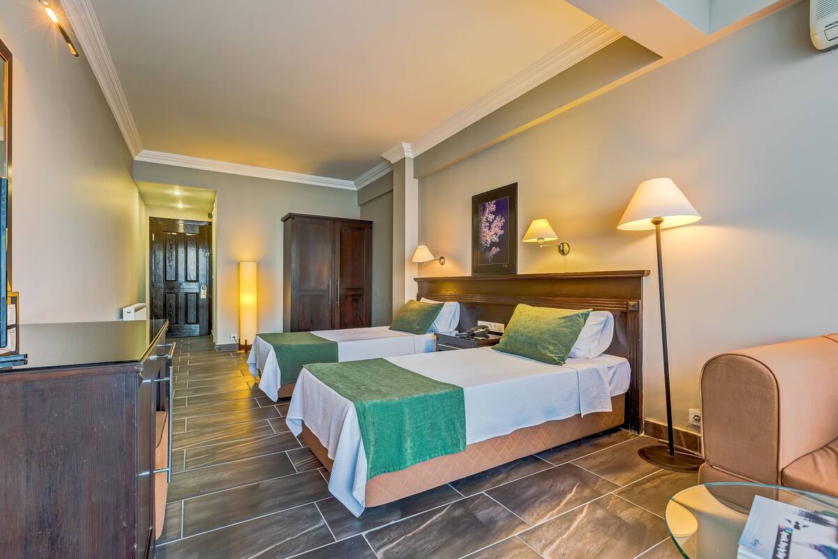 A hotel room with two beds, a desk, a chair, and a lamp.
