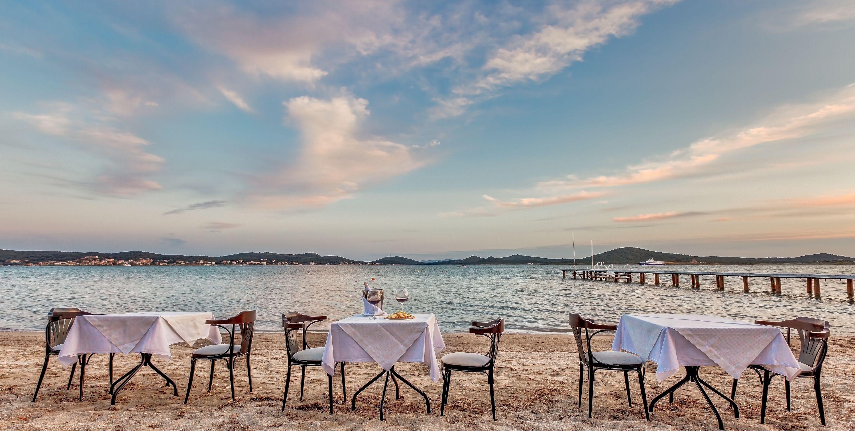 Ein Essbereich am Strand mit Tischen und Stühlen, die auf das Meer ausgerichtet sind, eine Pier, die sich ins Wasser erstreckt, und eine Küstenlinie in der Ferne mit Hügeln.
