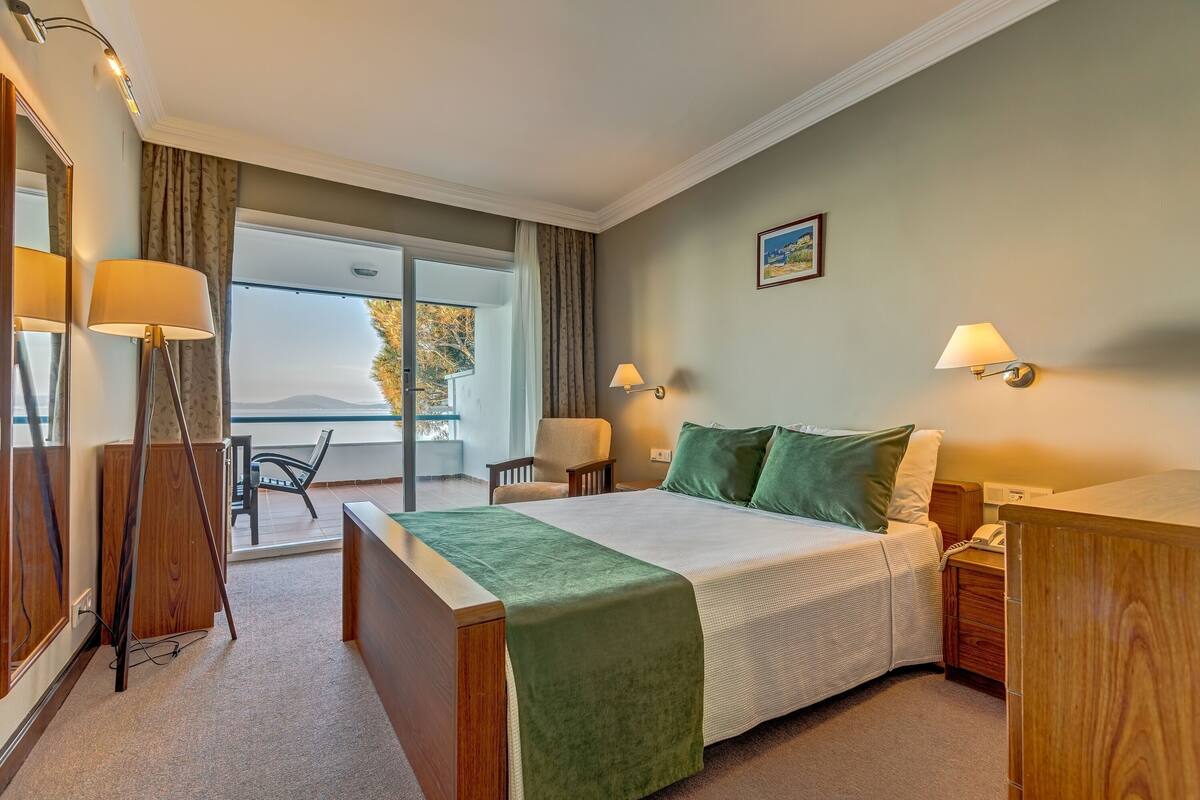 A bedroom with a bed, bedside tables, a desk, and a chair. There is a window with curtains and a view of the outdoors.