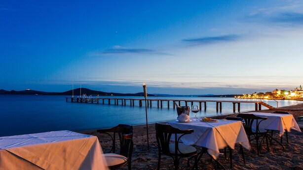 Een restaurant aan zee met tafels en stoelen op het strand, met uitzicht op een rustig wateroppervlak en een pier die zich in de verte uitstrekt.