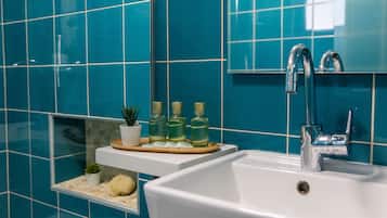 Uma casa de banho com uma pia branca, um espelho e azulejos verde-água. Há três frascos verdes num tabuleiro, uma pequena planta num vaso e uma toalha branca.