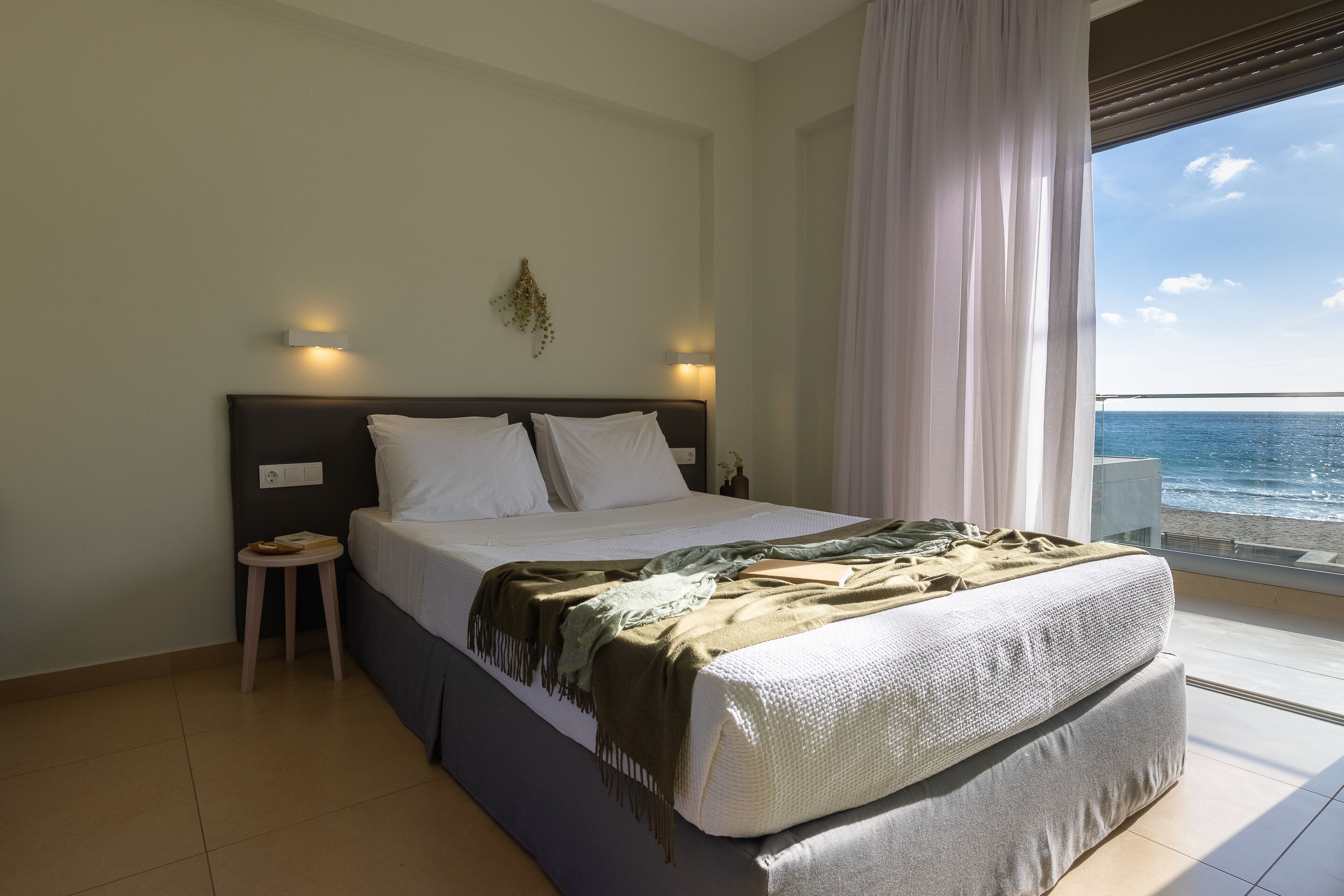 Quarto com uma cama grande, um banquinho pequeno, uma mesinha de cabeceira e vista para o mar.