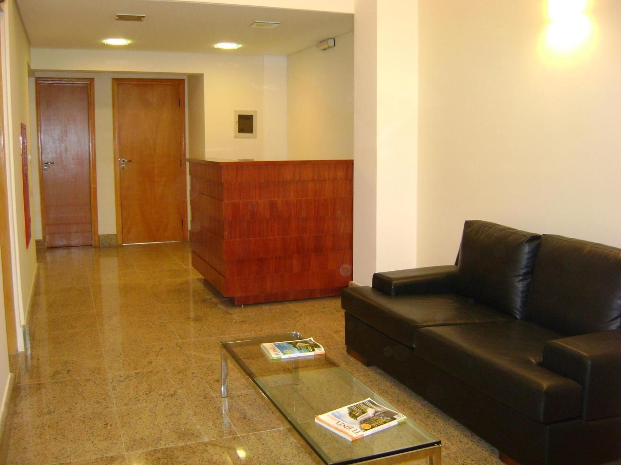Uma área de recepção com balcão de madeira, um sofá de couro preto e uma mesa de centro de vidro com revistas.