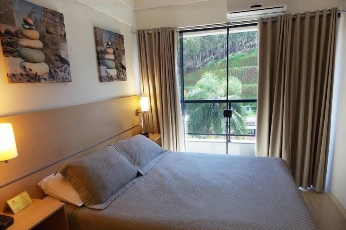 Habitación de hotel con una cama grande, una mesita de noche, una lámpara y un ventanal con cortinas.
