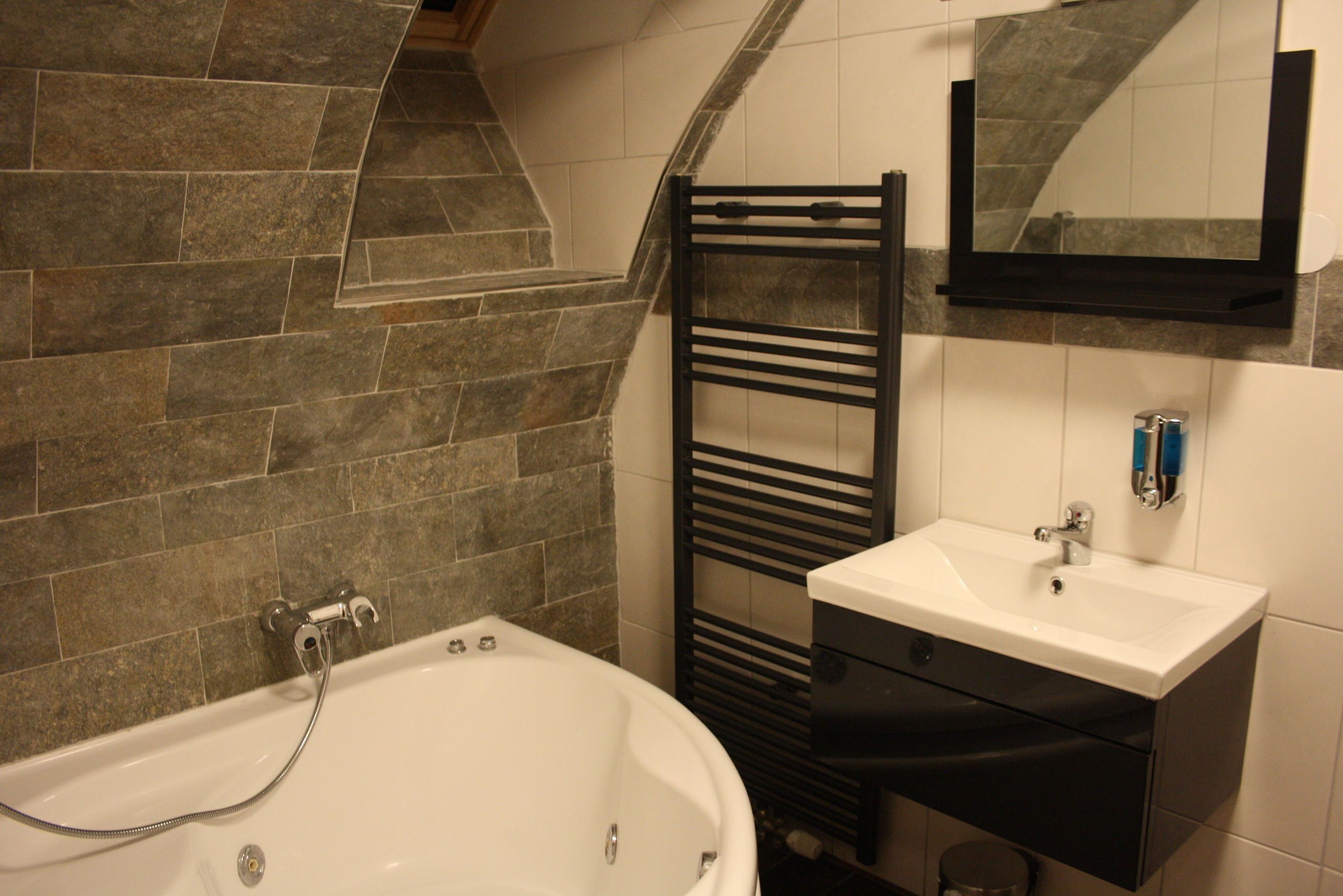 Une salle de bain avec un mur recouvert de carreaux de pierre, une baignoire blanche, un porte-serviettes noir, un lavabo blanc et un miroir au-dessus du lavabo.