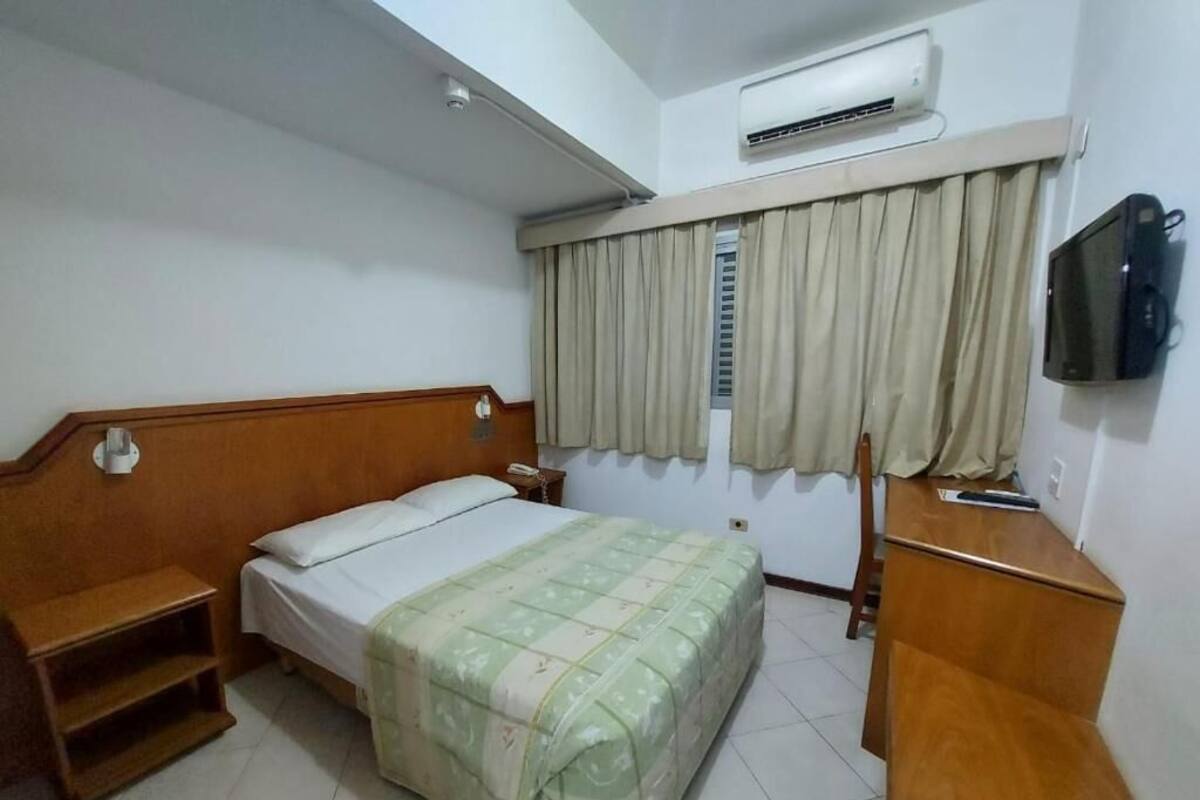 A hotel room with a bed, a wooden nightstand, a TV, and an air conditioning unit.