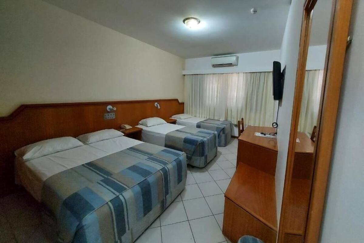A hotel room with three beds, a wooden desk, a mirror, and an air conditioning unit.
