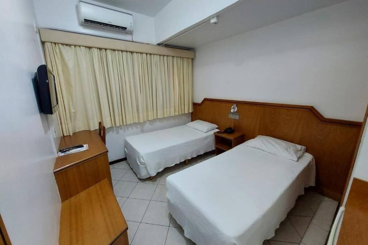 A hotel room with two single beds, a wooden desk, a chair, a nightstand with a lamp, and an air conditioning unit.