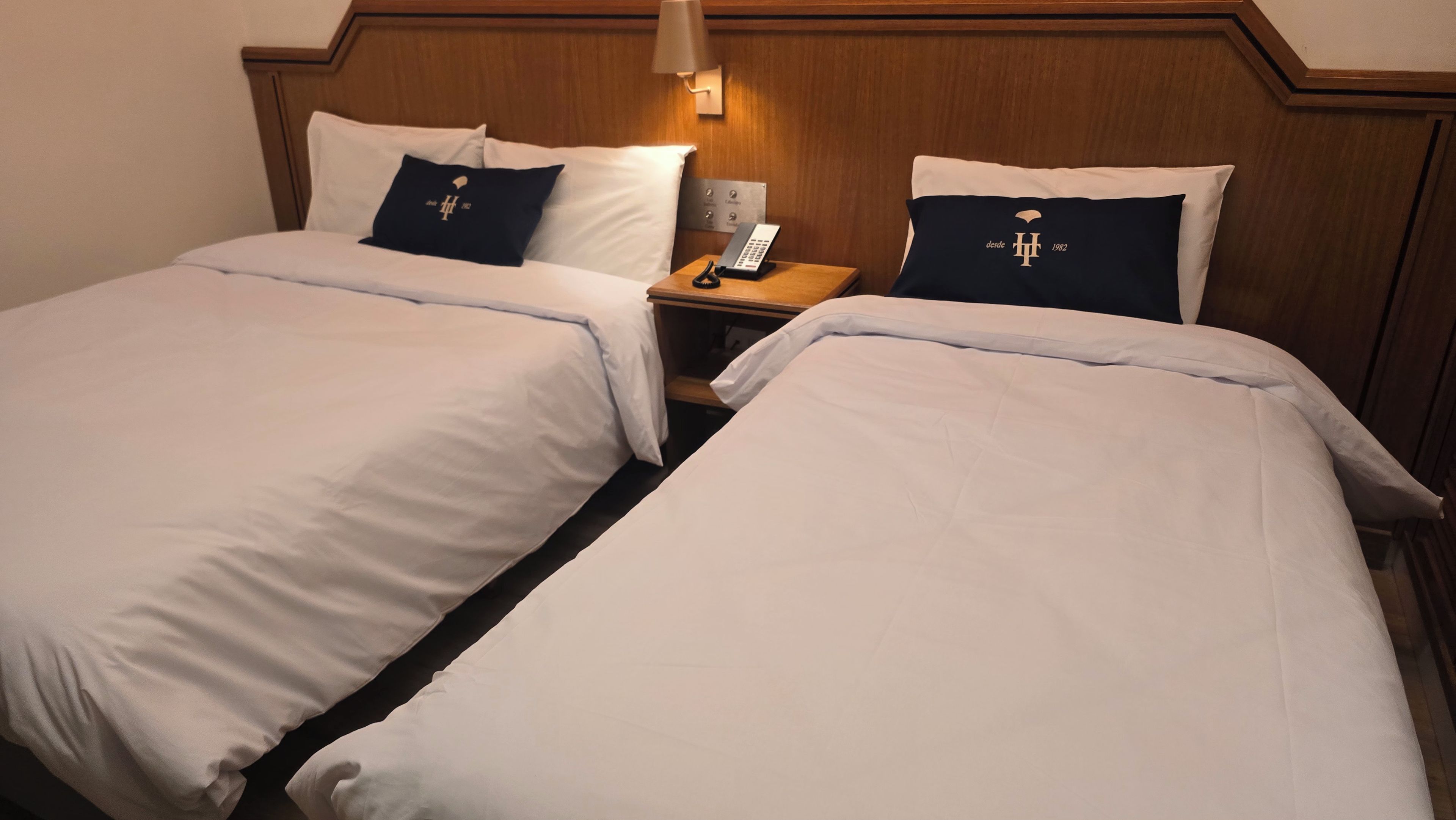 A hotel room with two beds, a nightstand with a phone, and a wall-mounted lamp.
