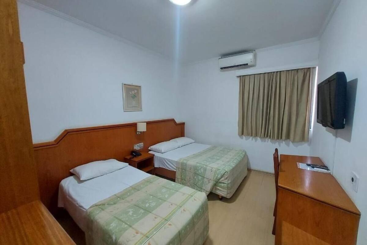 A hotel room with two beds, a wooden headboard, a nightstand, a desk, a television, and an air conditioning unit.