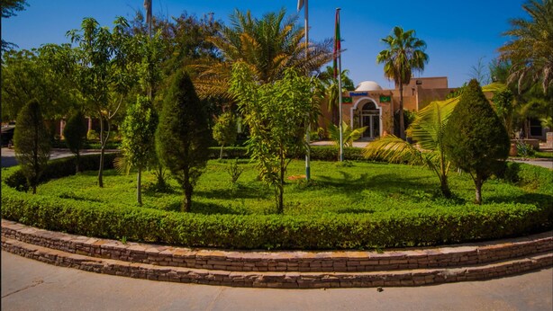 A landscaped garden with a building featuring a dome and flagpoles. - Monotel Dar El Barka (Nouakchott)