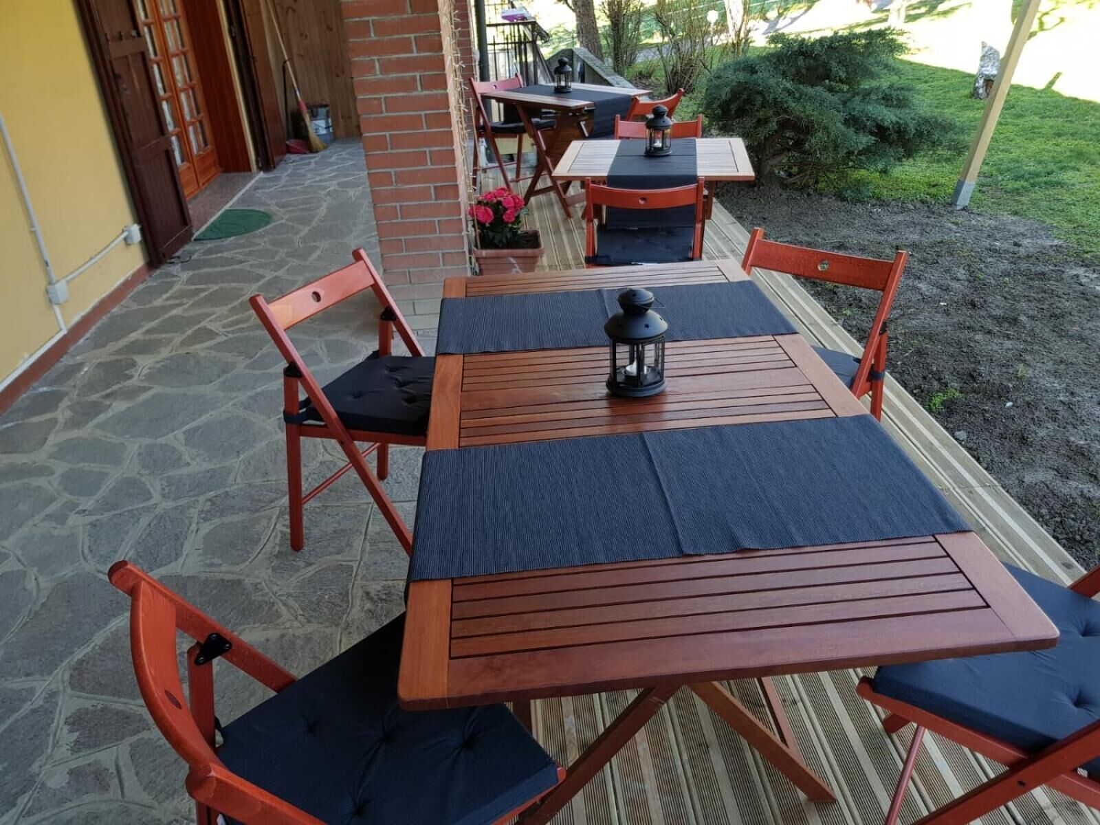 Eine Terrasse im Freien mit Holztischen und -stühlen, einige davon mit Kissen. Auf den Tischen stehen Blumentöpfe und ein Laternenlicht.