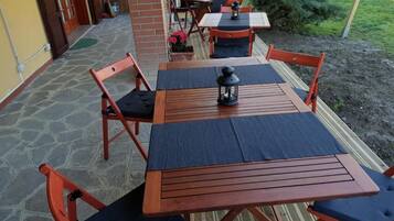 Eine Terrasse im Freien mit Holztischen und -stühlen, einige davon mit Kissen. Auf den Tischen stehen Blumentöpfe und ein Laternenlicht.