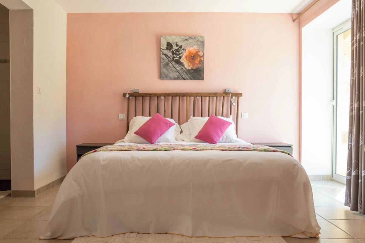 A bedroom with a bed, pillows, a nightstand, a wall-mounted artwork, and a window with curtains.