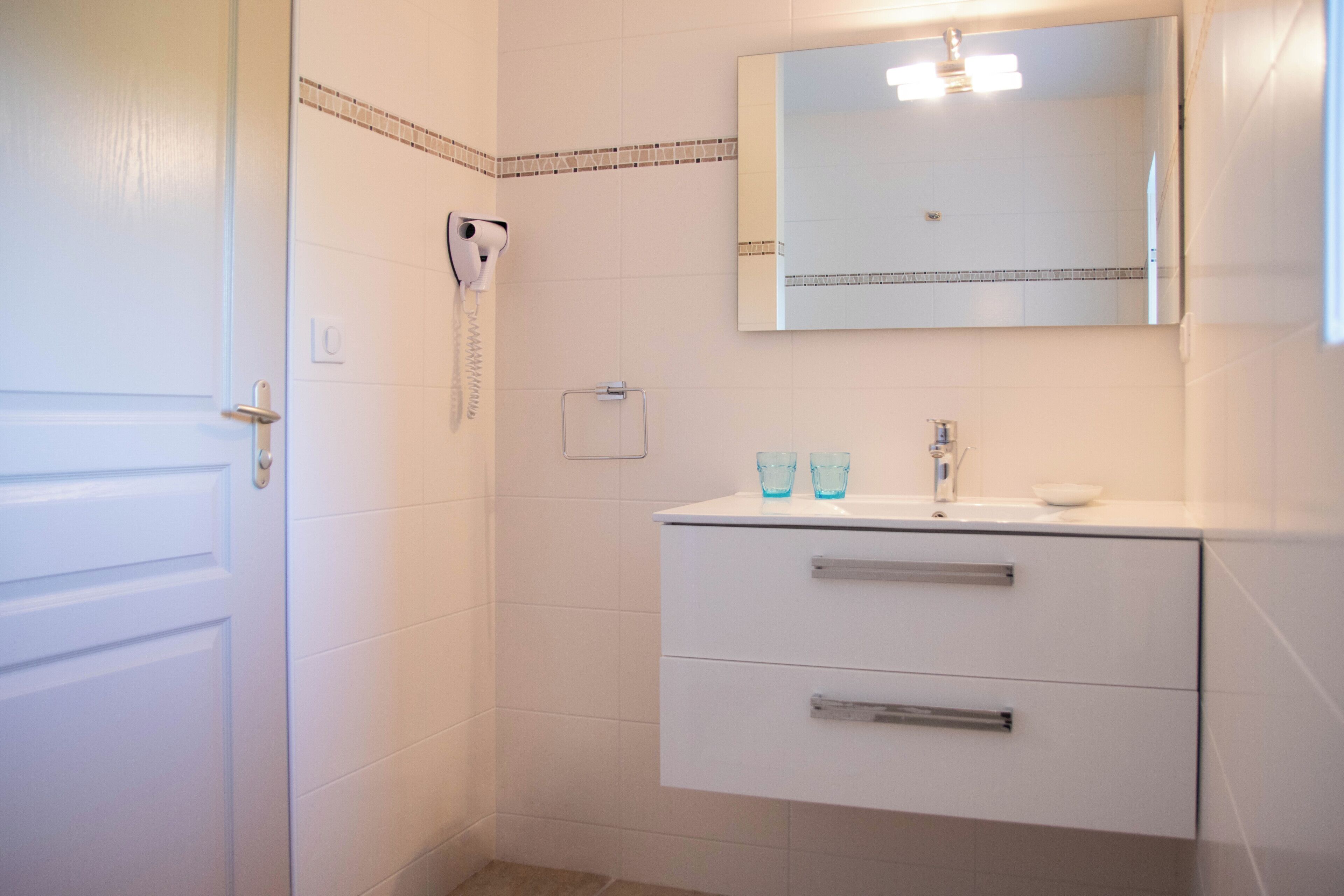 Un baño con puerta blanca, tocador blanco con dos cajones, espejo y zona de ducha con cabezal de ducha de mano.