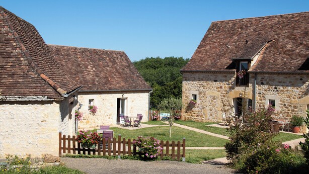 A stone building with a tiled roof, a wooden fence, and a small garden with flowers and chairs. - Domaine de Janis - Spa de nage (Le Vigan)