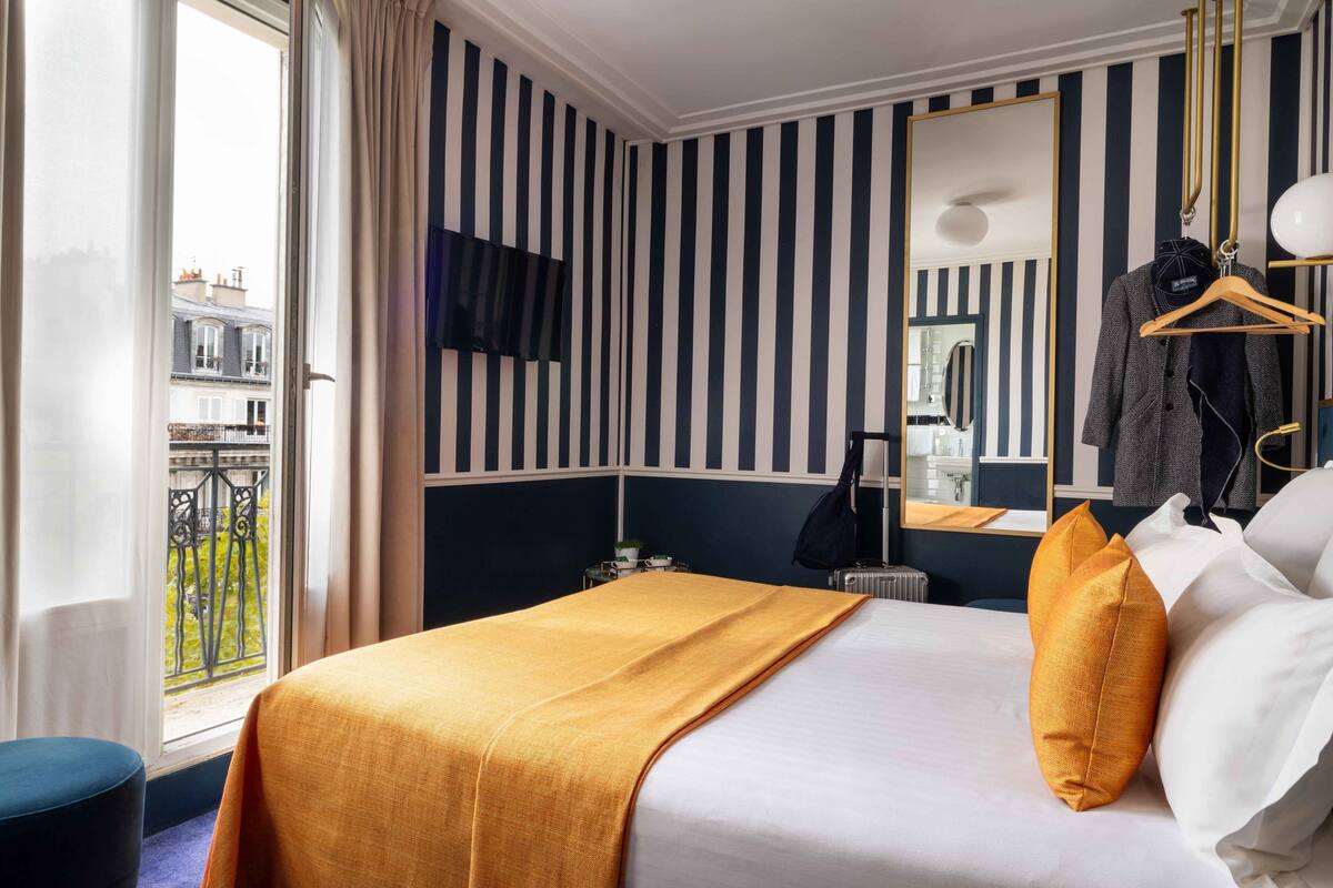 A hotel room with a large bed, a window with curtains, a striped wall, a mirror, and a hanging coat rack.