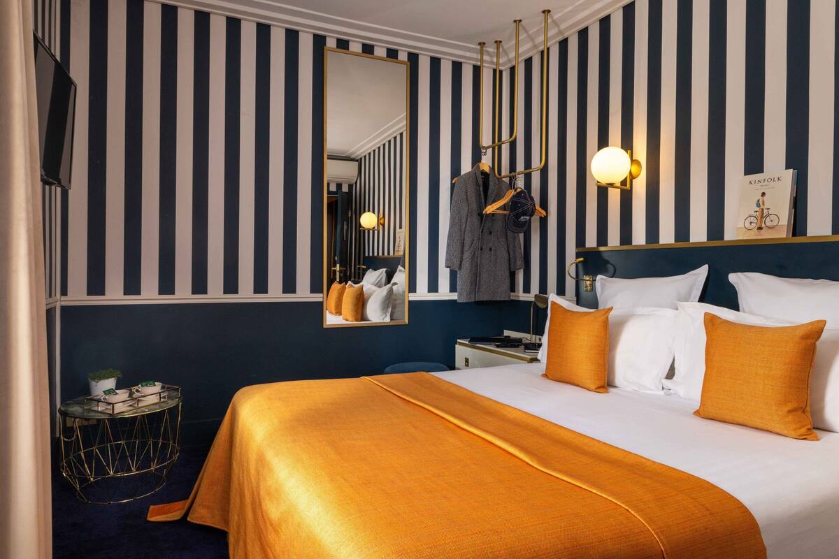 A hotel room with a striped wall, a large bed with white and orange bedding, a mirror, a coat rack, and a small side table with a plant.