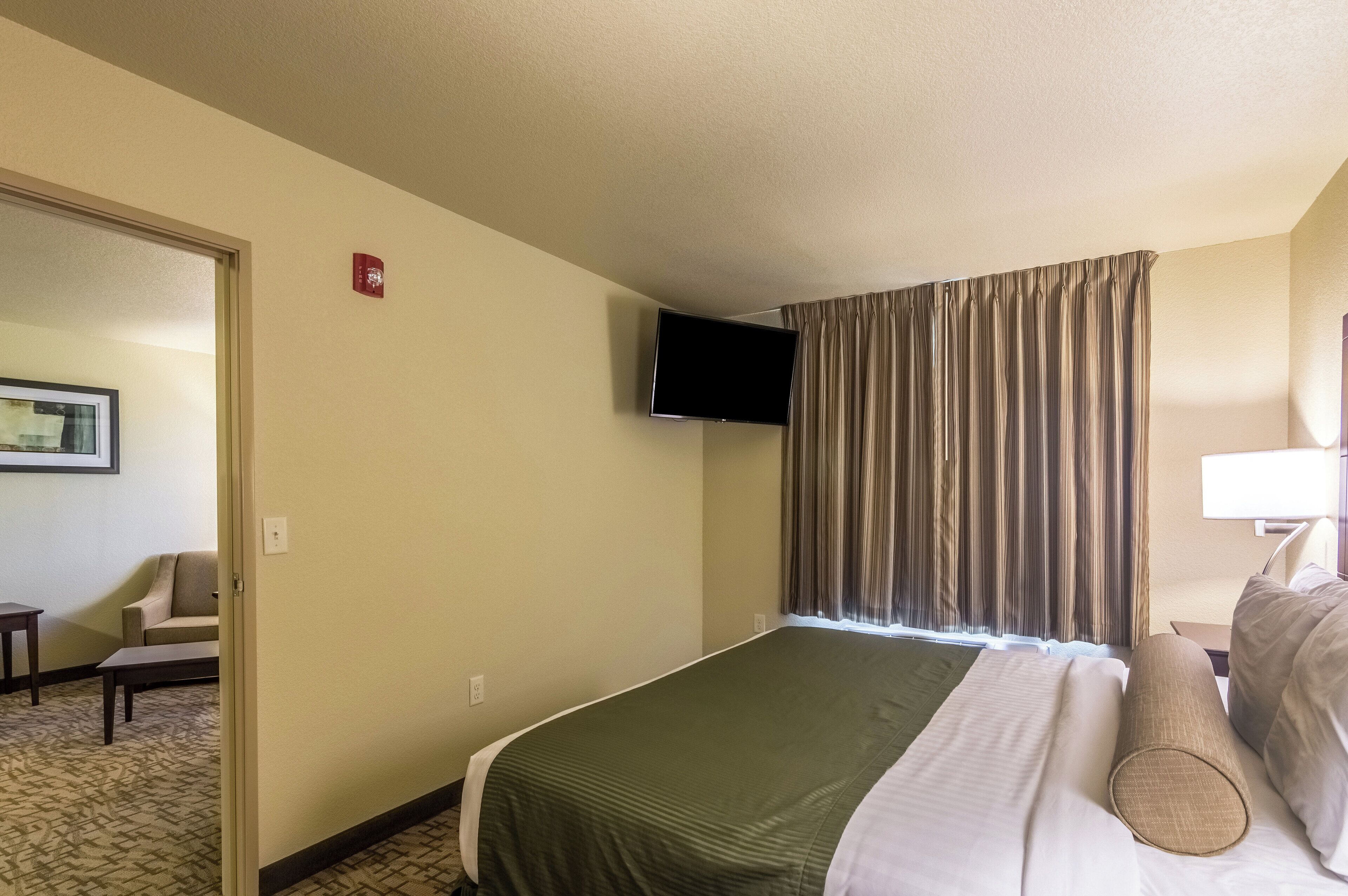 A hotel room with a bed, a television mounted on the wall, a window with curtains, a chair, and a small table.